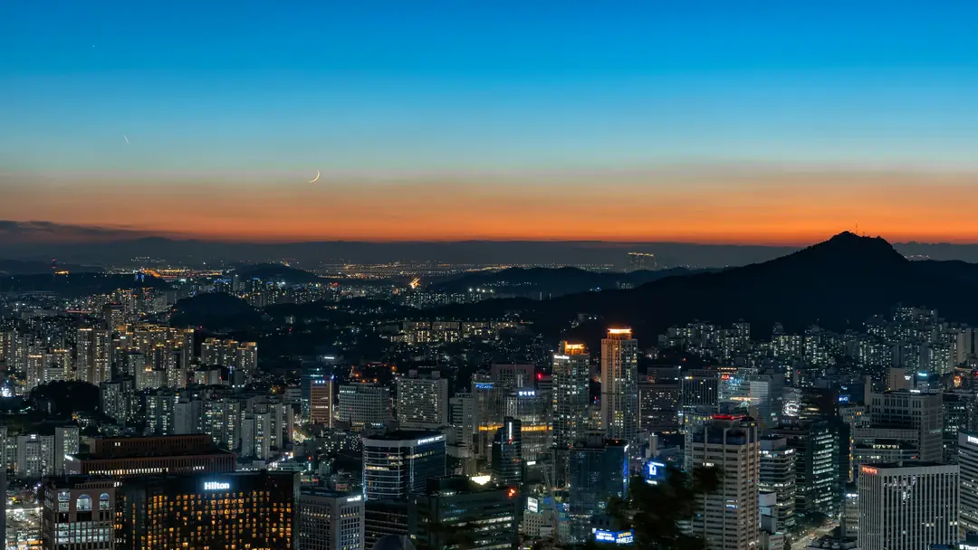 The Seoul skyline lit up at dusk under a crescent moon