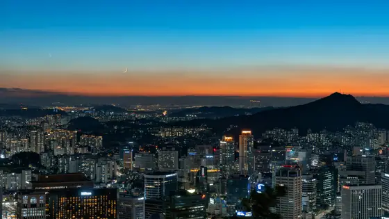 The Seoul skyline lit up at dusk under a crescent moon